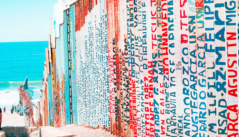 The border wall on the Mexican side. The wall extends into a bright blue ocean, and there are names painted on the metal bars in blue, red, and black.