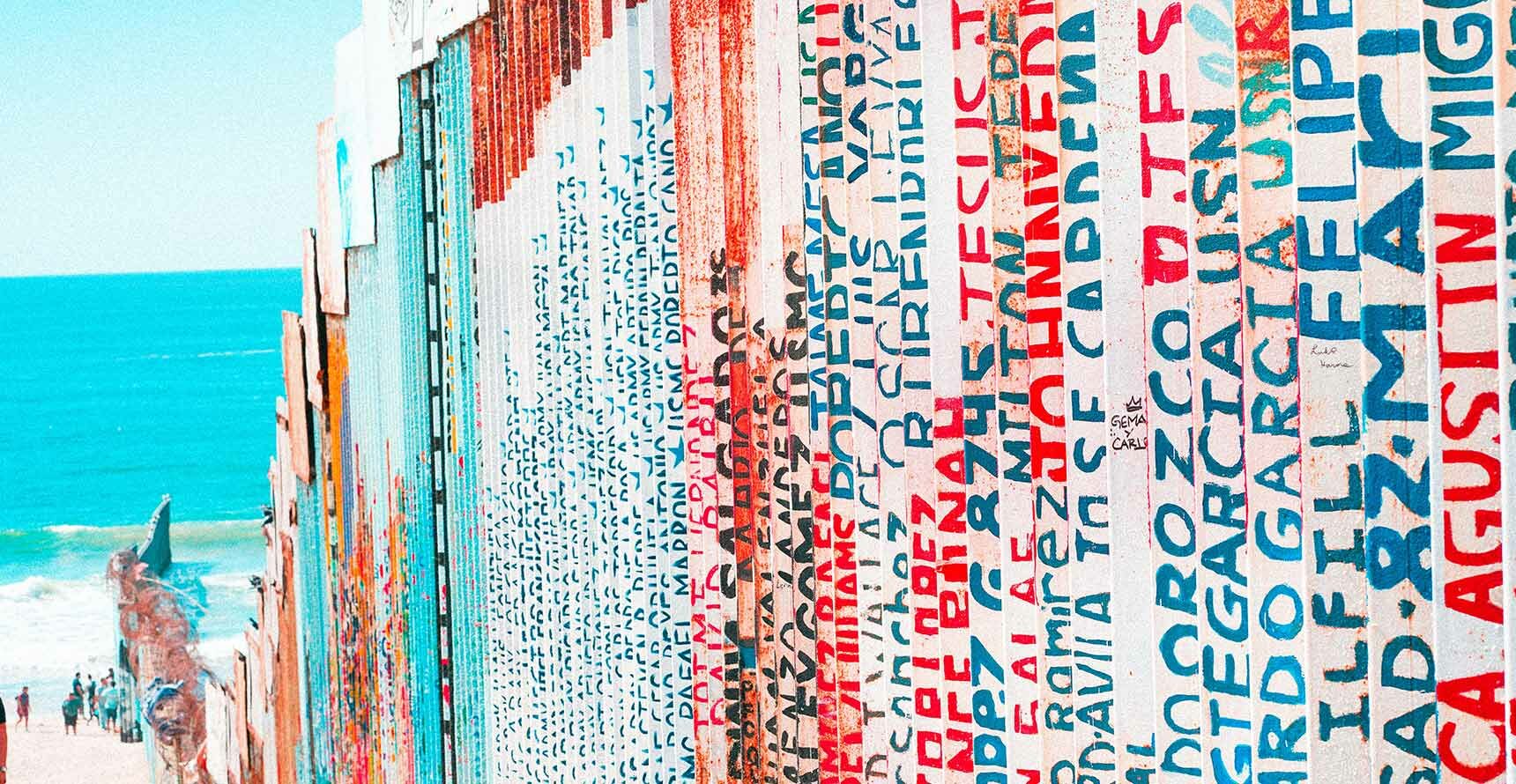 The border wall on the Mexican side. The wall extends into a bright blue ocean, and there are names painted on the metal bars in blue, red, and black.