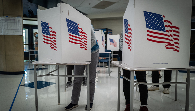 Two white voting booths with American flags on them. Two people are hidden behind the voting booth.