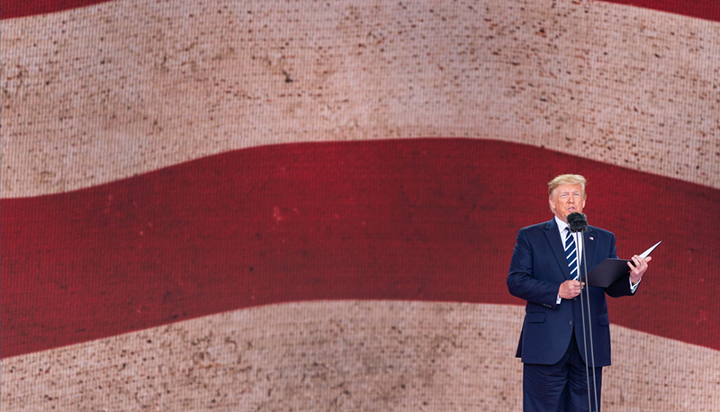 President Donald Trump stands in front of a large wall depicting the American flag