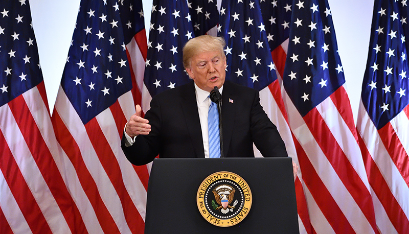 President Donald Trump speaks at a podium in front of a backdrop of several U.S. flags